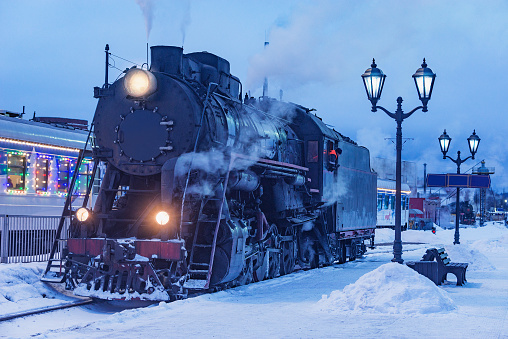 Retro steam locomotive stands by the station platform at winter evening.