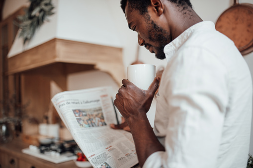 Candid millennial authentic diverse portrait of african american man relax time in neutrals tones kitchen. Brown skin male drink tea at breakfast domestic life and morning routine reading newspaper