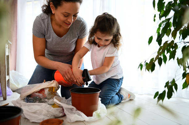 Cute baby girl watering soil in pots while planting houseplants with her mom in home balcony stock photo