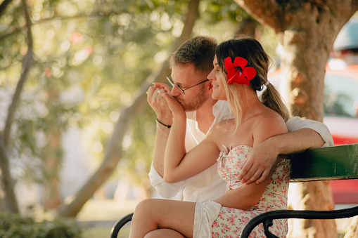Young man kissing his beautiful girfliend's hand while they are sitting on bench on vacation. The girl has red Hibiscus flower in her hair.