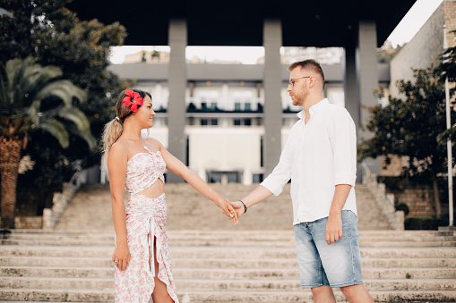 Young man and woman holding hands and looking at each other, at the background stairs to hotel.