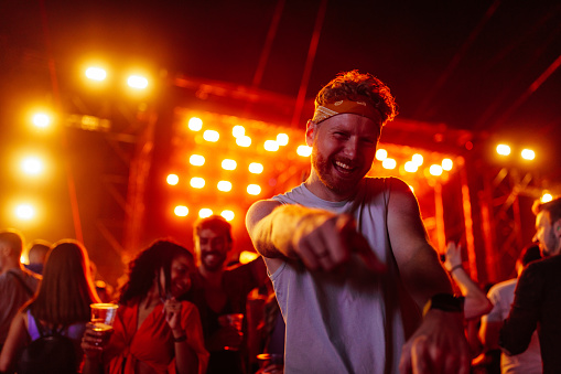 A young adult Caucasian man is dancing at a music festival on a beautiful summer night with the stage vibrantly lit behind him.