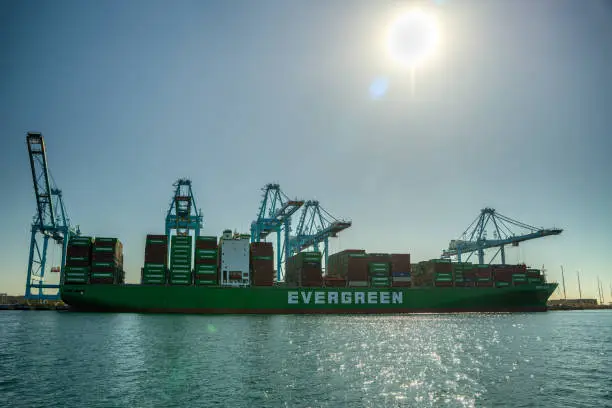 Container transportation is now the most popular way of cargo movement and a critical pillar of the global economy, Drone shot of a cargo ship sitting in dock in the Port of Long Beach, with crane booms lowering into position to unload the containers.