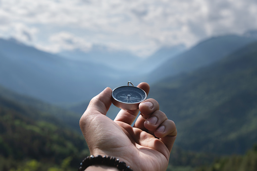 Magnetic compass in the palm of a male hand against the backdrop of a mountain range in the clouds in the summer outdoors, travel, first-person view