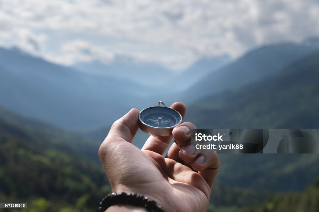 Magnetic compass in the palm of a male hand against the backdrop of a mountain range in the clouds in the summer outdoors, travel, first-person view Magnetic compass in the palm of a male hand against the backdrop of a mountain range in the clouds in the summer outdoors, travel, first-person view. Navigational Compass Stock Photo