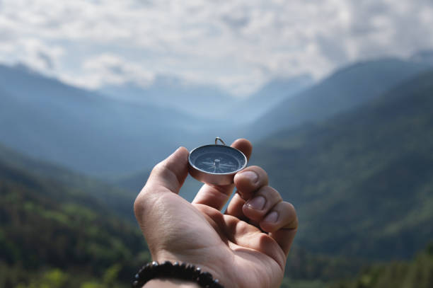 Magnetic compass in the palm of a male hand against the backdrop of a mountain range in the clouds in the summer outdoors, travel, first-person view stock photo