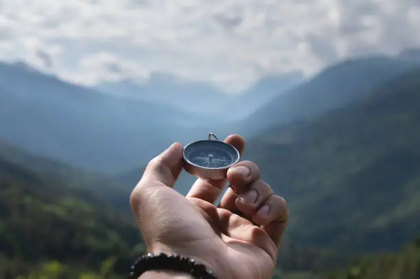 Photo of Magnetic compass in the palm of a male hand against the backdrop of a mountain range in the clouds in the summer outdoors, travel, first-person view