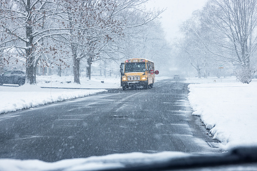 A yellow school bus with flashing light stop signs extended with flashing lights is stopped during a windy blizzard snow storm on a suburban street to let school children get off the bus at their home driveway.