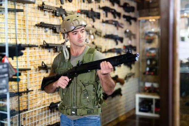 Caucasian man wearing armor vest and helmet while choosing machine gun in airsoft shop.