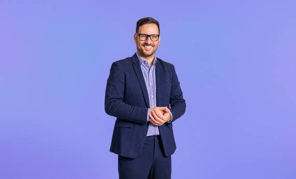 smiling elegant mid adult male manager dressed in blue suit clasping his hands while standing confidently isolated over blue background - mans suit imagens e fotografias de stock