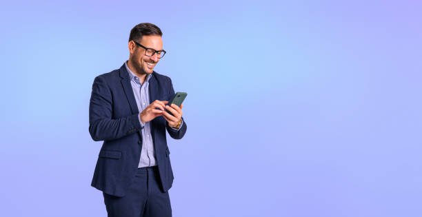 smiling mid adult businessman dressed in elegant formalwear texting online over smart phone and standing isolated on blue background - clothing well dressed business waist up imagens e fotografias de stock