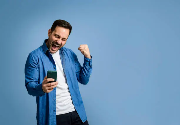 Photo of Handsome mid adult man dressed in denim shirt screaming and cheerfully pumping fist while reading good news over smart phone on blue background