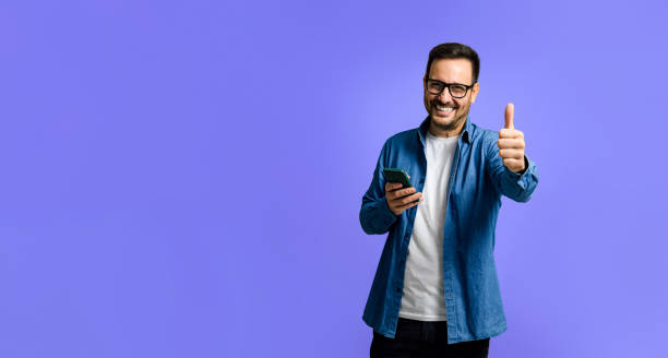 retrato do homem jovem adulto alegre na camisa jeans usando o telefone inteligente e mostrando o sinal de polegares para cima enquanto está isolado sobre o fundo azul - mid adult men smart casual white happiness - fotografias e filmes do acervo