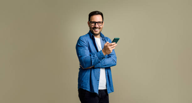 istock Smiling young adult man text messaging over smart phone and looking at camera while posing confidently against isolated gray background 1473323348