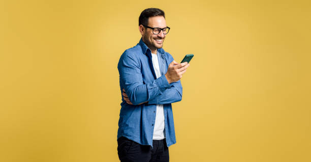 beau jeune homme adulte souriant vêtu d’une chemise en jean bavardant sur un téléphone intelligent et se tenant isolé sur fond jaune - isolated happiness carefree one person photos et images de collection