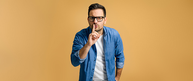 Serious mid adult man dressed in blue denim shirt with finger on lips showing silence gesture while standing isolated on orange background