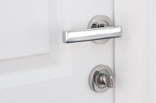 Close up view of opened modern white door with nickel handle and lock. Grey wall near the entrance. Minimalistic interior design. Copy space.