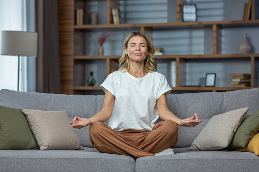 Senior woman at home resting meditating with eyes closed sitting on sofa in living room, housewife mature blonde in lotus pose smiling.