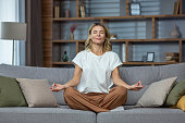 Senior woman at home resting meditating with eyes closed sitting on sofa in living room, housewife mature blonde in lotus pose smiling