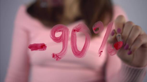 若い女性は、ガラスの割引からマイナス90%を引き出しています。ピンクのセーターを着た女の子がブラシと赤い絵の具で描きます。クローズ アップ。4キロメートル競走 - number 20 percentage sign number glass ストックフォトと画像