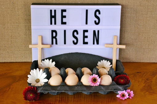 A Lightbox with the words HE IS RISEN with a wooden religious cross, eggs and flowers for a religion or easter background.