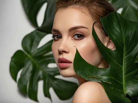 Closeup face of young beautiful woman with a healthy clean skin. Beautiful white girl with big green leaves. Beauty and spa treatment concept. Pretty woman with natural makeup and plant near face