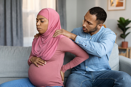Loving husband making massage to his pregnant black muslim wife at home, expectant islamic woman in hijab suffering from lower back pain or prenatal contractions while sitting on couch in living room
