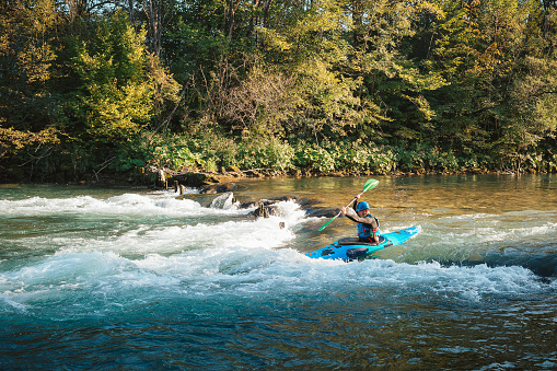 Young teenager cruising down whitewater rapids in a blue kayak, beautiful river nature on a sunny summer day, handheld shot.