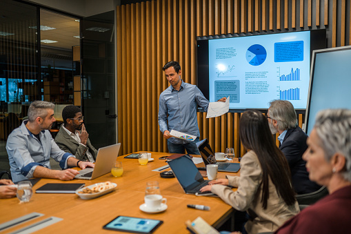 Mid adult Asian businessman standing in front of a big screen in a modern board room holding a meeting and updating board members with past and future business results.