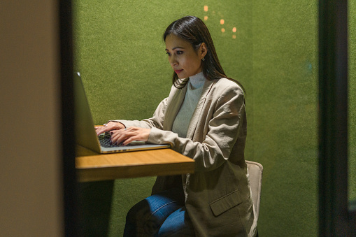 Asian businesswoman in early 40s using a small guest office in a busy corporate building. She is using a laptop while preparing for a business meeting.