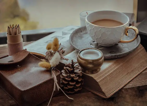 Morning refreshing with Hot coffee in ceramic cup 
served with Fresh milk on old wooden worktable by the window view with old book, colored pencils, small handbag leather and dried flowers. Selective focus.
