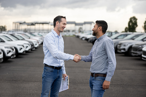 Happy salesman handshaking with a client after buying a car at the dealership - car ownership concepts