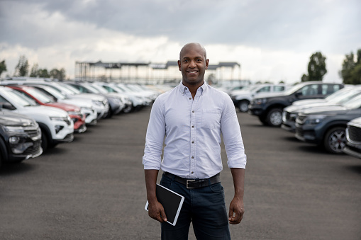 African American salesman working at a car dealership selling cars and looking at the camera smiling - automobile industry concepts