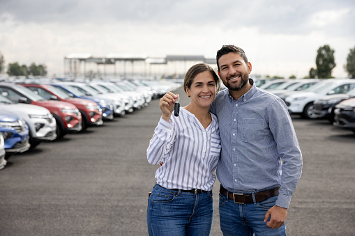 Portrait of a happy Latin American couple holding the keys of their new car at the dealership - car ownership concepts