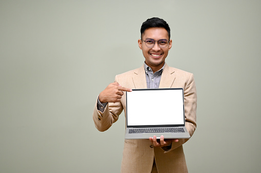 Happy and smiling adult Asian businessman in formal business suit holding his laptop, showing laptop blank screen, pointing his finger at laptop screen, standing against green background.