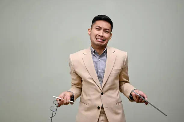 Reluctant and unpleasant millennial Asian businessman or male office worker in formal business suit holding stylus pen and tablet, standing against a green background. unhappy, disgusting, unwilling