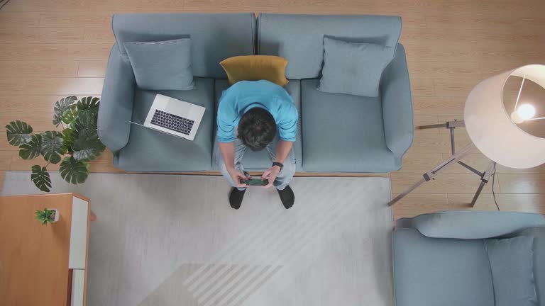 Top View Zoom Out Shot Of A Man Playing Game On Smartphone While Sitting On The Sofa Works On A Laptop At Home