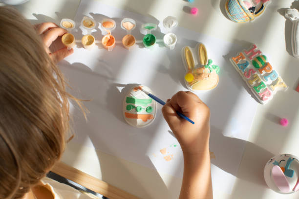istock A cute little girl is sitting at a table and doing Easter crafts, coloring Easter figurines. Creativity and hobbies. 1473410451