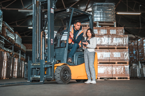 Handsome young Asian female supervisor working with a male warehouse employee. He is sitting on a forklift. She is scrolling on a digital tablet.