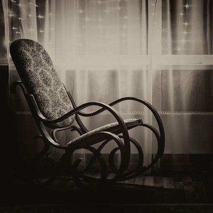 A rocking chair stands by the window in the evening light. Black and white sepia.