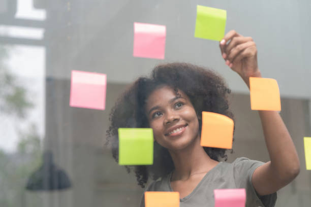 istock Young smiley attractive, businesswoman using sticky notes on a glass wall to write a strategic business plan to develop and grow to success. 1473417125
