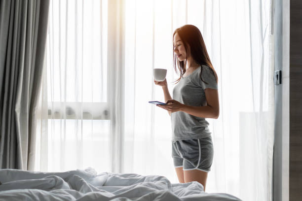 istock Young beautiful brunette hair woman in pajamas drinking coffee and using mobile phone while standing near the window in bedroom in the morning. Copy space 1473420321