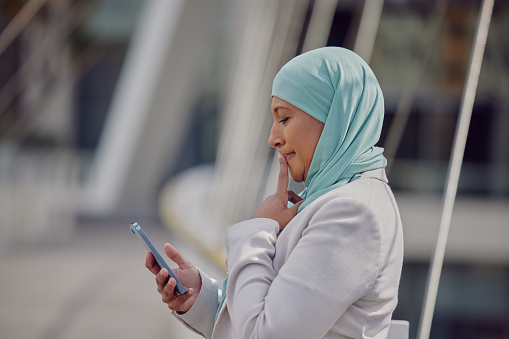 Muslim woman, phone and thinking in the city for business research, communication or social media. Thoughtful Islamic female employee with hijab on smartphone wondering in search on app in Dubai
