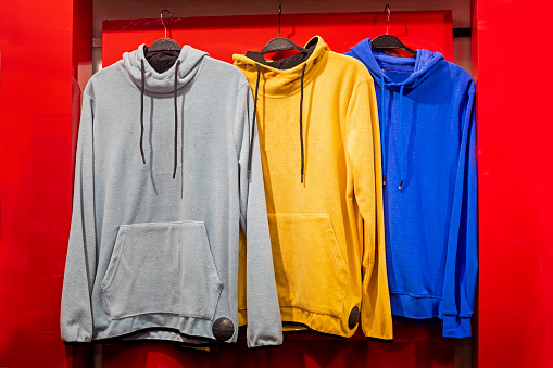 Multicolored hoodie hang on hangers against the background of shelves with clothes in the store.