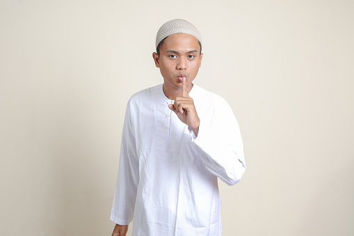 Portrait of attractive Asian muslim man in white shirt whispering malicious talk conversation, hand on mouth telling secret rumor. Isolated image on gray background