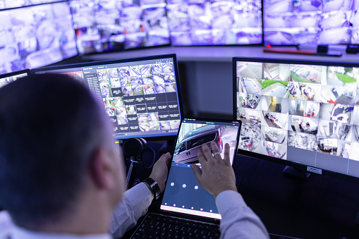 Mature adult man working in surveillance, security and guarding agency and looking at many monitors in front of him and using control panel on tablet.
