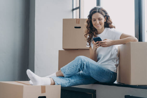 istock Happy spanish woman is sitting on windowsill texting on phone and unpacking boxes. 1473443497