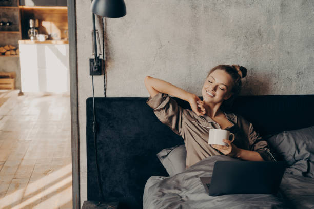 istock Happy beautiful female with closed eyes drinking coffee in bed before remote work at home 1473445278