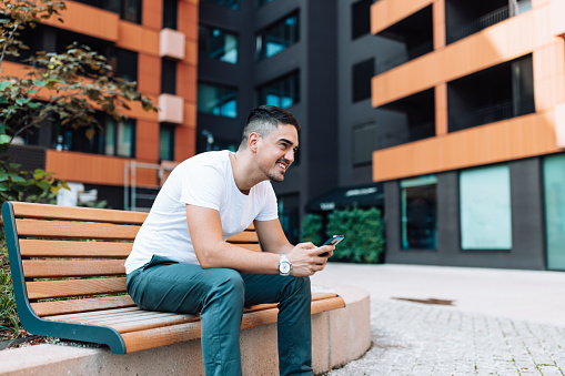A young Caucasian man is looking up from his phone, while sitting on a bench outside.