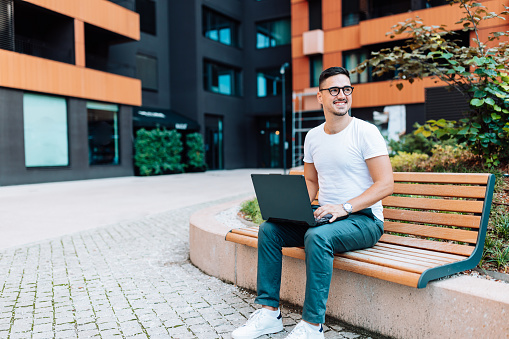 A young Caucasian man wearing eyeglasses is looking up from his laptop with a smile, while sitting on a bench.